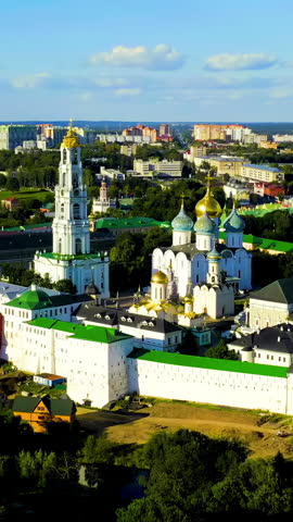 Vertical video. Sergiev Posad, Russia. The Trinity Lavra of St. Sergius, a UNESCO World Heritage Site and the largest operating male monastery of the Russian Orthodox Church, dominates the city center with its striking ensemble of white-walled fortifications, golden onion domes, and an 88-meter baroque bell tower. Founded in 1337 by St. Sergius of Radonezh, the complex grew over five centuries into a remarkable fusion of traditional Russian and Western European architectural traditions. The aerial footage captures the Lavra from an elevated perspective, revealing the full scale of the fortified compound with its green rooftops, multiple cathedral domes catching the warm sunset light, and the surrounding urban landscape of Sergiev Posad. The golden hour illumination highlights the contrast between the pristine white facades, the gilded cupolas, and the deep green roofs, while the modern city stretches toward the horizon beyond the historic walls. This active monastery complex, which withstood a 16-month siege during the Time of Troubles, remains a major pilgrimage destination and a defining landmark of the Golden Ring of Russia. Aerial View, Point of interest. Rich colors