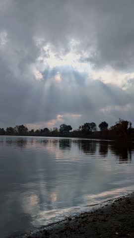 A vertical view of Sun rays break through heavy clouds over a calm river, casting dramatic light and reflections on the water