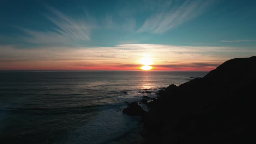 A coastal sunset over calm ocean water, with dark cliffs framing the horizon under streaked clouds and warm evening light