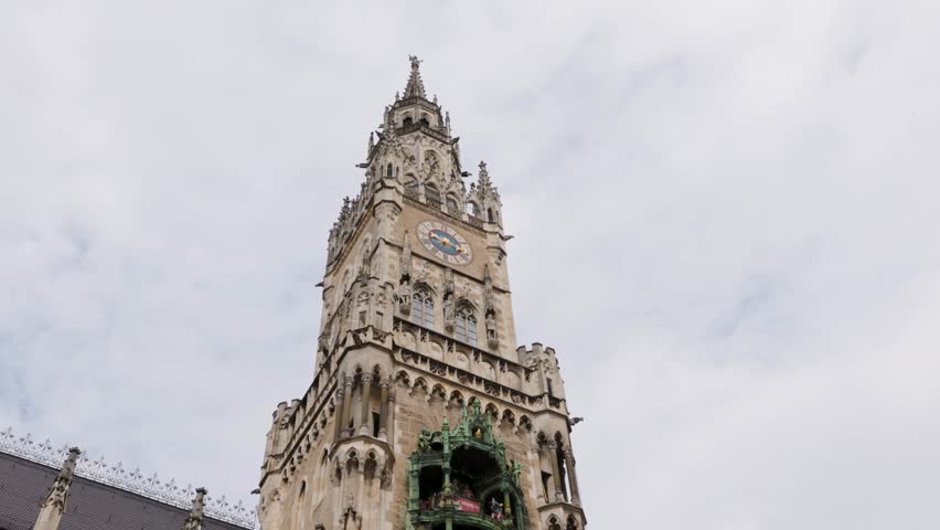 A detailed view of a Gothic Revival clock tower featuring ornate stonework and multiple colorful vertical flags hanging from the adjoining building.