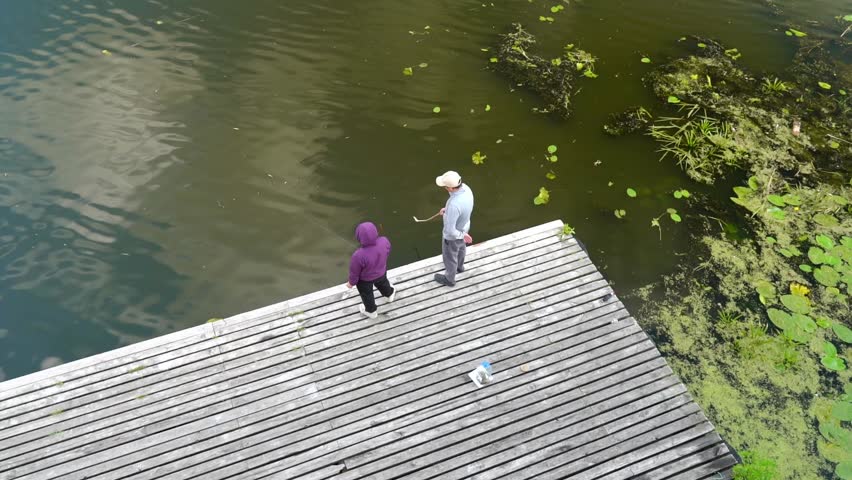 Two people stand on a wooden dock by a lake with green plants in the water. They are fishing and enjoying the time together in a quiet spot.in Belarus, in the city of Pinsk, on the embankment of the Pina River, on July 19, 2026