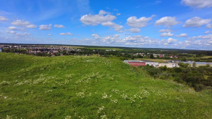 The photo shows a wide view of a green landscape with hills and clouds. Houses and buildings are visible in the distance under a blue sky. People can enjoy nature and outdoor recreation. A beautiful view of the town of Vawkavysk in Belarus from a Swedish mountain.