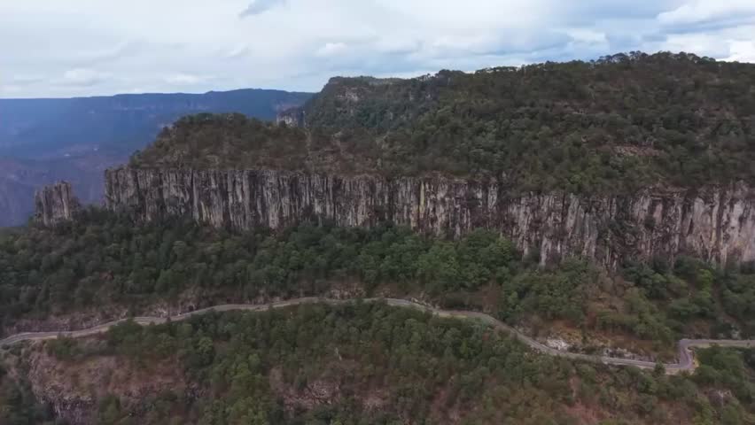 Wide aerial drone shot showcasing a scenic serpentine highway carving through dense green mountain forest, positioned right underneath impressive vertical rock formations under a soft cloudy sky.