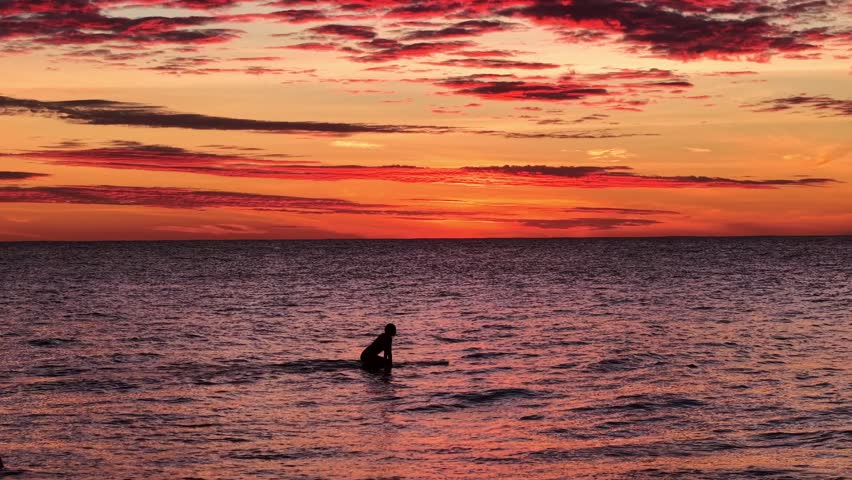 Silhouette of lone surfer sitting on surfboard waiting for waves in calm ocean water under vibrant red sunset sky.