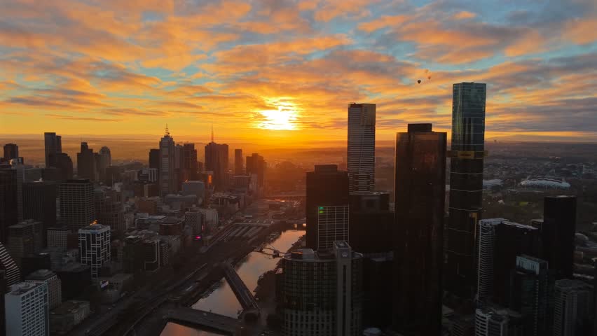 Aerial drone video over Melbourne CBD and Southbank at sunset, flying above the Yarra River with city skyscrapers, bridges and glowing golden clouds