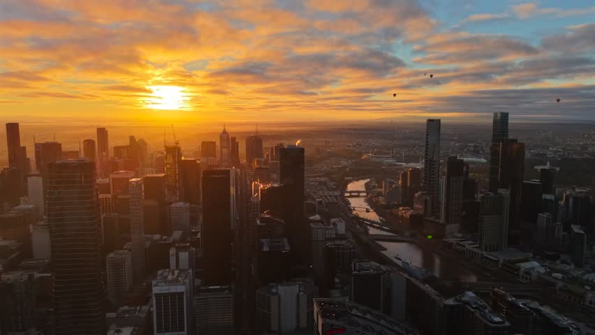 Aerial drone video flying right over Melbourne CBD at sunrise toward Southbank, with the Yarra River, skyscrapers, hot air balloons and glowing clouds