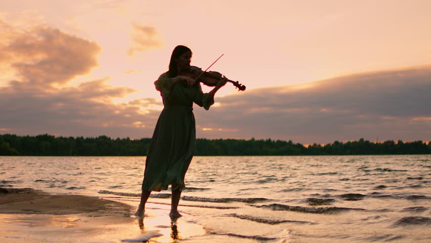 Silhouette of a young woman playing violin on a tranquil lakeshore at golden sunset, waves gently touching the sandy beach under dramatic colorful sky