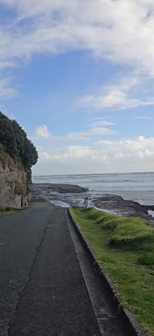 Scenic coastal walkway overlooking a rugged ocean shoreline with dramatic waves, lush green cliffs, and a wooden bench under a vibrant blue sky with scattered clouds. A peaceful seaside landscape capturing the natural beauty of New Zealand
