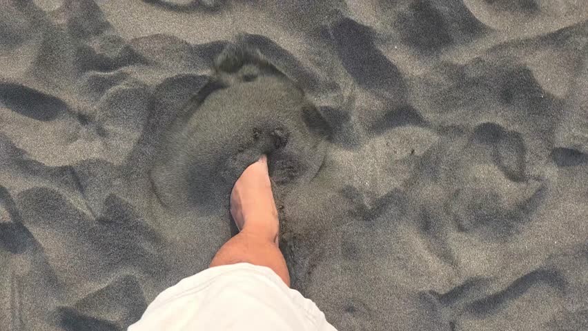 High angle view of bare feet walking on soft grey beach sand during a sunny day.