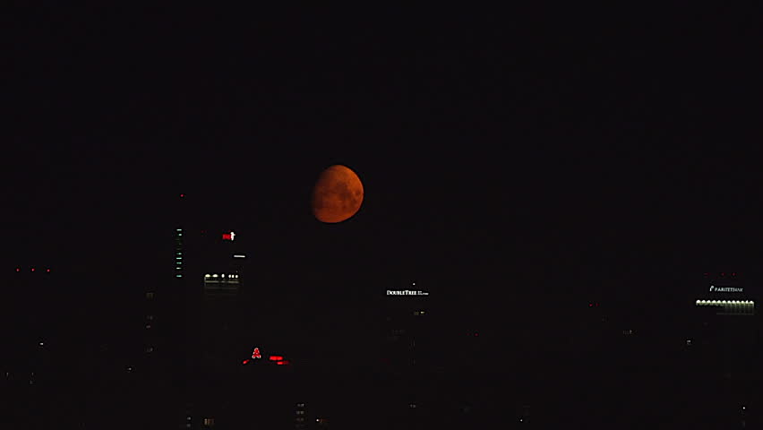 A dramatic cinematic shot of a massive, glowing blood red moon rising low over a modern city skyline at night. Hyper-detailed skyscrapers with glowing windows reflecting the crimson moonlight. Dark moody atmosphere, wispy night clouds passing in front of the moon, deep red and orange color palette. Shot on a telephoto lens, compression effect making the moon appear giant behind the buildings.