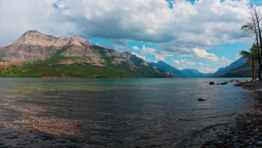 A wide-angle view of a clear mountain lake bordered by rocky shoreline and distant peaks, captured under dramatic clouds and soft daylight