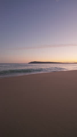 Sunset overlooking a beautiful sandy beach with waves washing into the shore, in the background you can see a landscape with a colourful orange and purple sky above. Located on the coast of Australia.