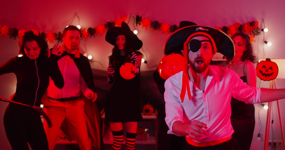 Joyful young man in pirate costume celebrating at Halloween party with friends. Group of people in spooky outfits dancing, having fun in decorated room with festive lights and pumpkin decorations.