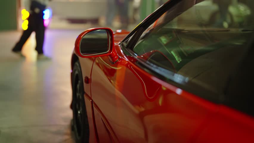 Close up view of a luxury red sports car parked in an underground garage with people walking past in the blurred background and colorful lights reflecting on the shiny surface of the car