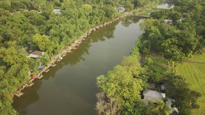 Drone shot moving in a semicircle from one side of the pond to the other, showing a community fishing event, gathered people, fishing stalls, shoreline activity, and a peaceful rural setting in Bangladesh.