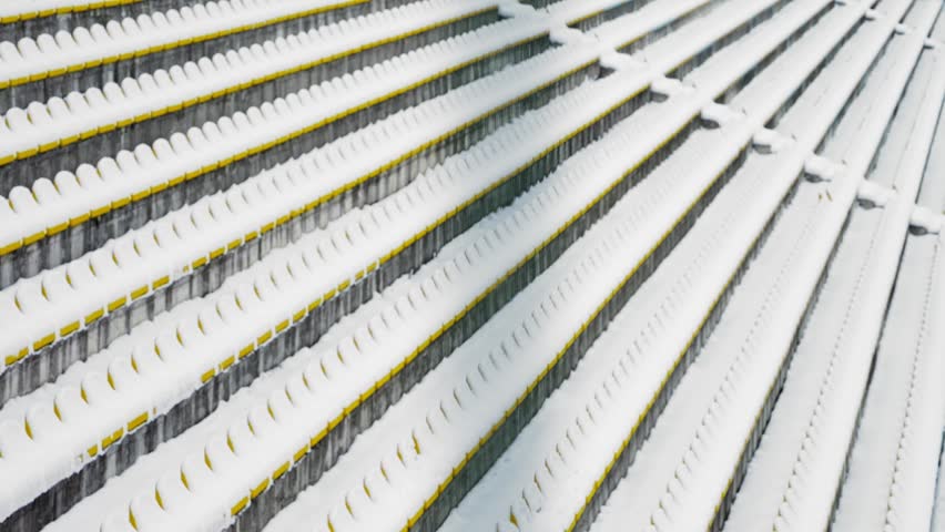 Fresh snow blankets rows of yellow plastic chairs inside an outdoor stadium. Winter conditions transform the sports venue into a calm architectural scene featuring clean lines, bright colors.