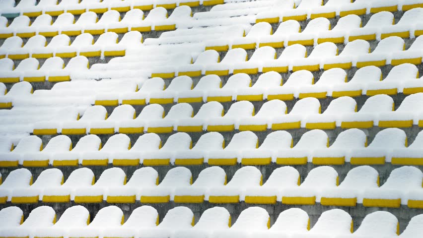 Fresh snow covering rows of yellow plastic seats in an outdoor stadium during winter. Peaceful architectural scene with colorful bleachers, snowy weather, sports venue, and seasonal atmosphere.