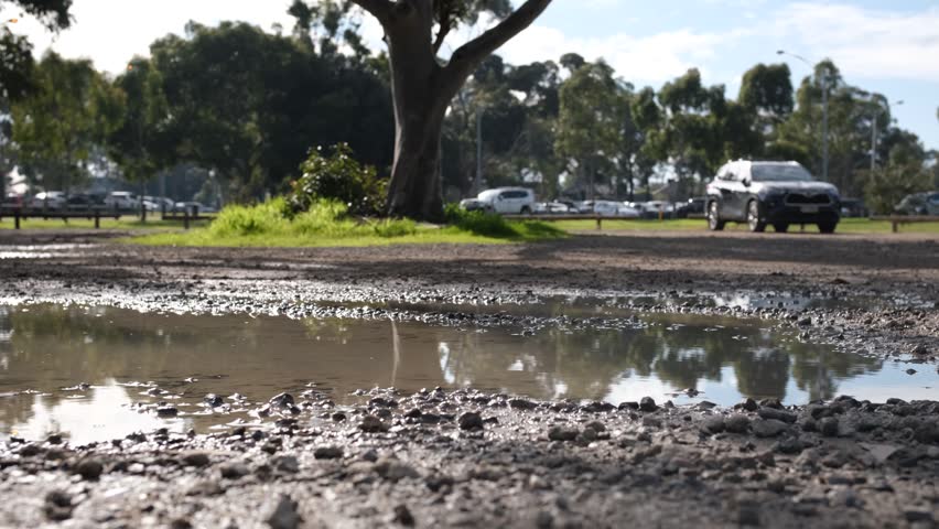 A muddy puddle covers a gravel unsealed car park in a suburban park. Infrastructure neglect, council maintenance issues, and wet weather conditions in Melbourne Australia.