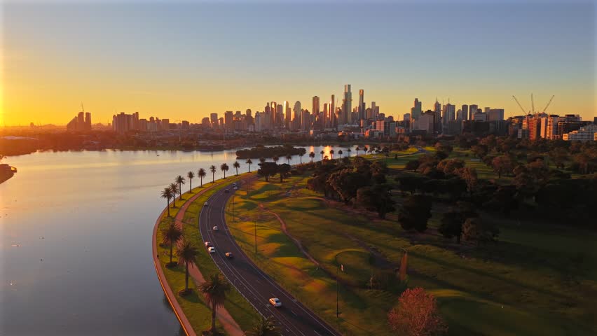 Low aerial drone video flying toward Melbourne CBD at sunset over Albert Park Lake, showing palm-lined roads, golf course, Southbank and golden city skyline