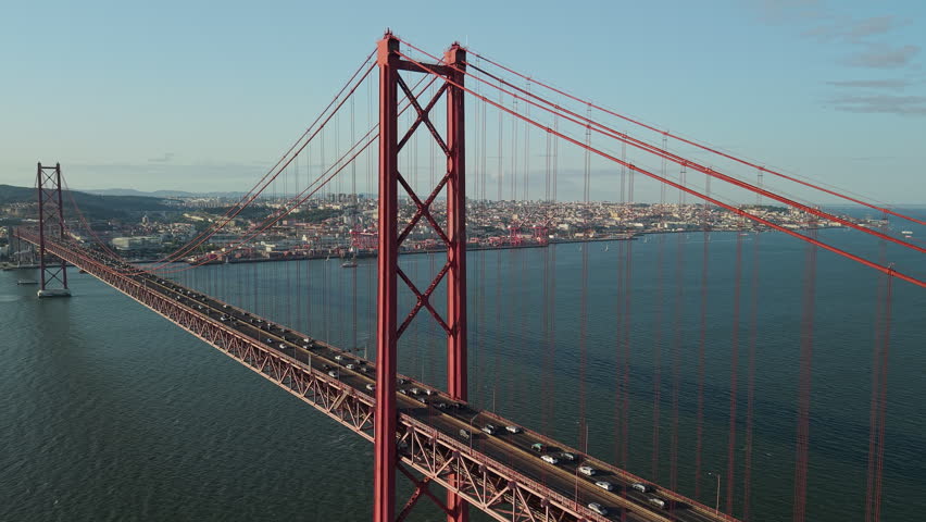 Aerial panoramic view of the red 25 de Abril suspension bridge spanning the Tagus River with traffic, Lisbon skyline, harbor, and port cranes in the background.