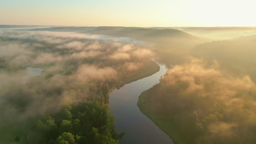 Aerial view of winding river cutting through dense forest, wrapped in morning mist under warm golden sunrise light.