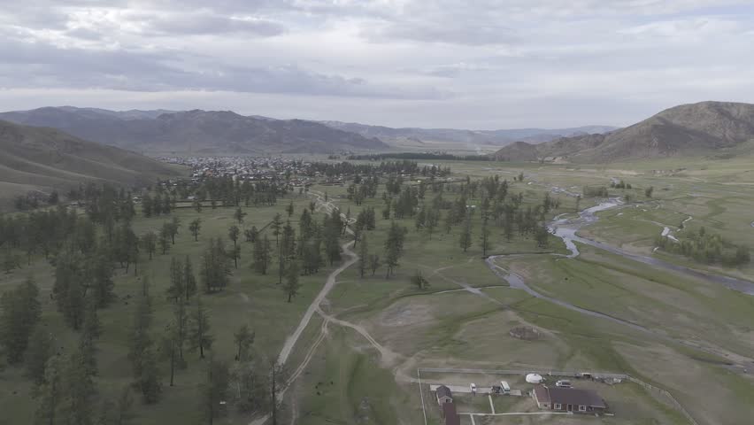 A drone view of a winding river flowing through the Mongolian steppe beside a small rural camp, scattered trees, and wide grasslands under daylight