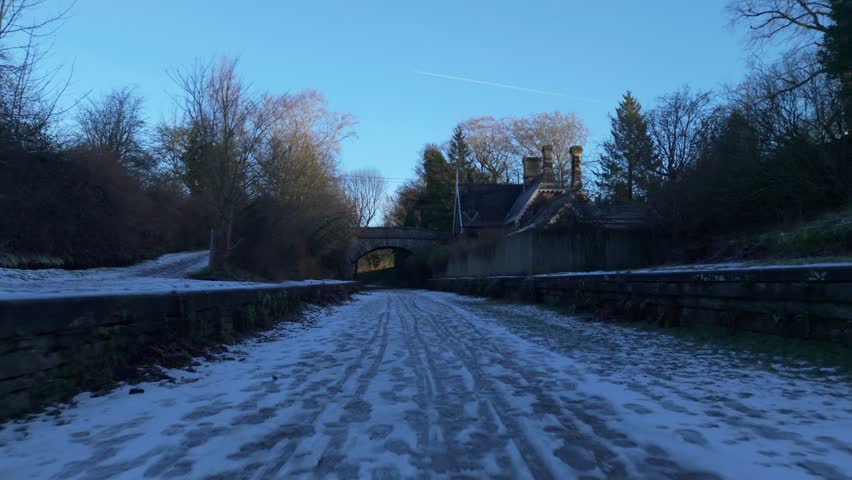 A snowy path leads under an old stone bridge beside winter trees and a quiet house in cold light