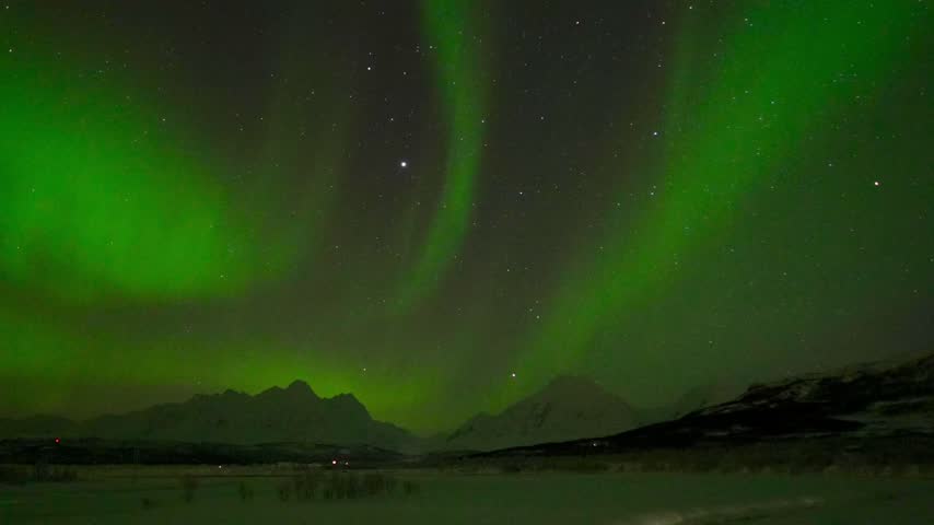 A Timelapse shot of green aurora lights glowing across a starry night sky above dark mountains