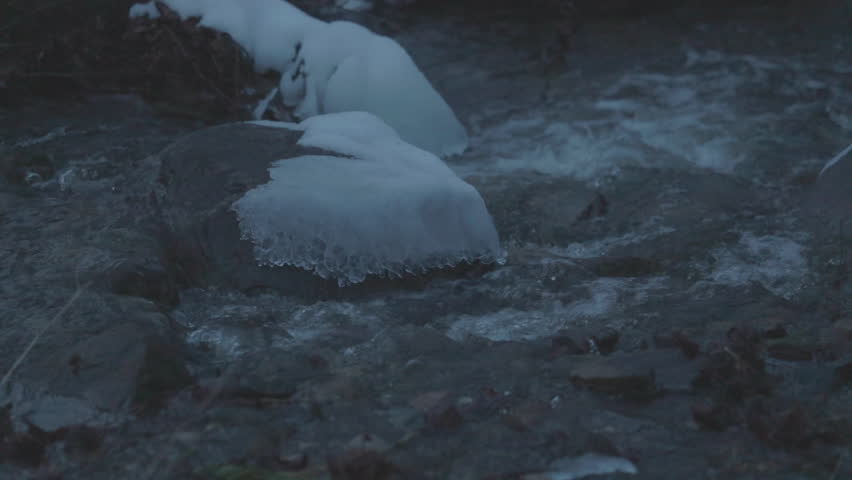 A winter stream detail showing water flowing around snow covered rocks, creating a cold and peaceful nature scene under soft twilight light