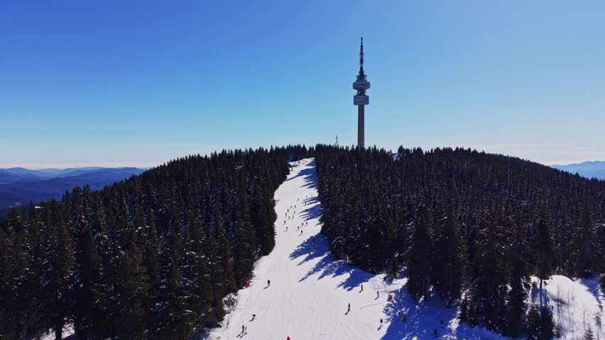 People ski down a snow-covered slope in the Carpathian mountains of Bulgaria while others enjoy the winter landscape. A tall tower stands on the hill under a clear blue sky.