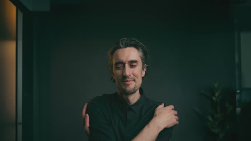 Close-up portrait of an adult man in a black shirt making contemplative gestures indoors with soft lighting and minimal decor.