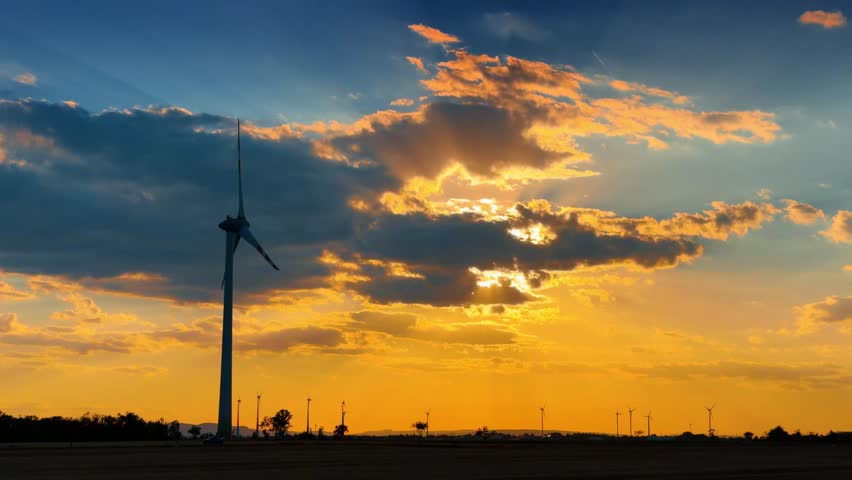 Wind turbine silhouette against dramatic sunset clouds. Sun rays break through dark cloud formations behind a tall power generator tower.