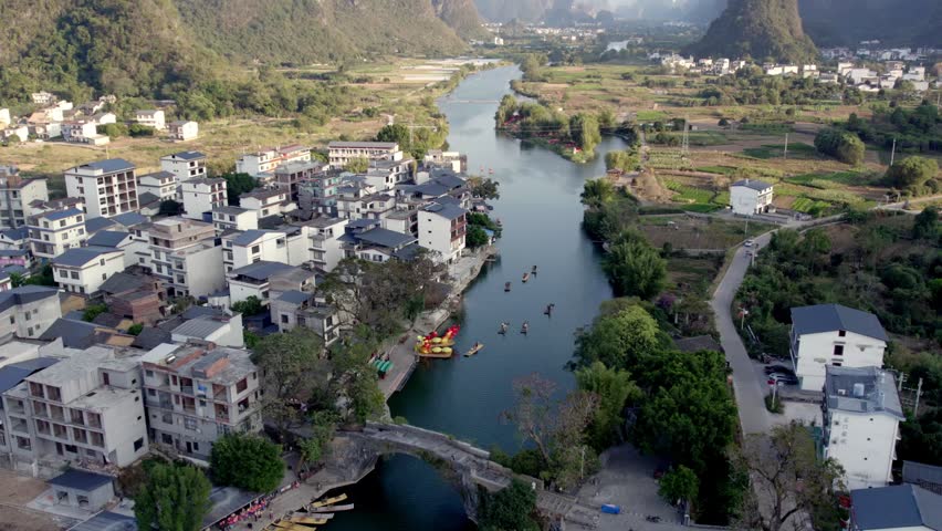 An aerial view of a riverside settlement with a stone arch bridge, flanked by karst mountains and farmlands under daylight.