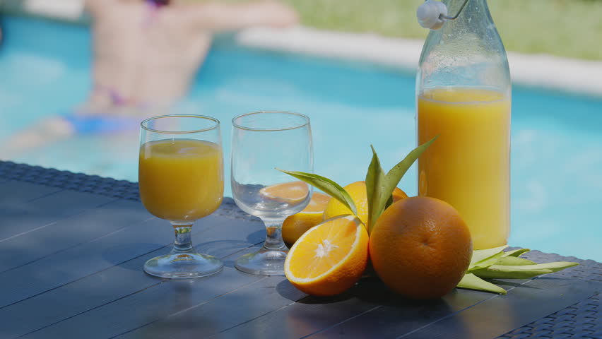 A man pours orange juice into a glass on a poolside table with blurred background of two young women sitting on the edge of the pool.