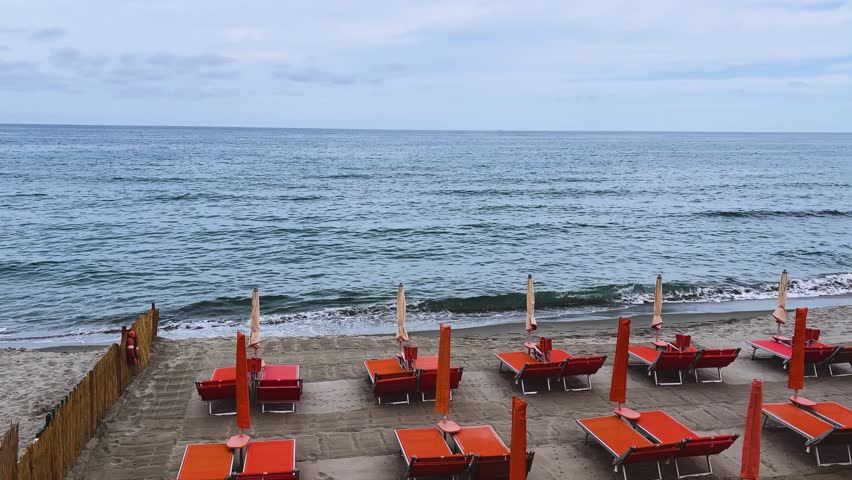 Orange loungers and closed umbrellas line a quiet beach on Italy’s Ligurian coast, with gentle waves rolling beneath a cloudy sky.