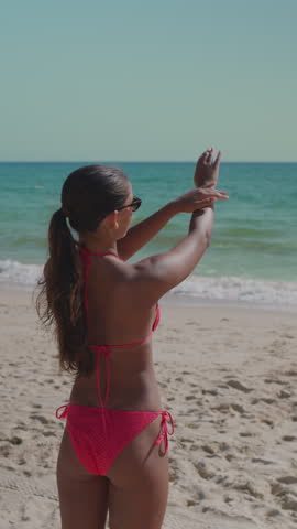 Young attractive teen woman with dark long hair applying sunscreen lotion on her hand on sandy beach near ocean, medium shot.
