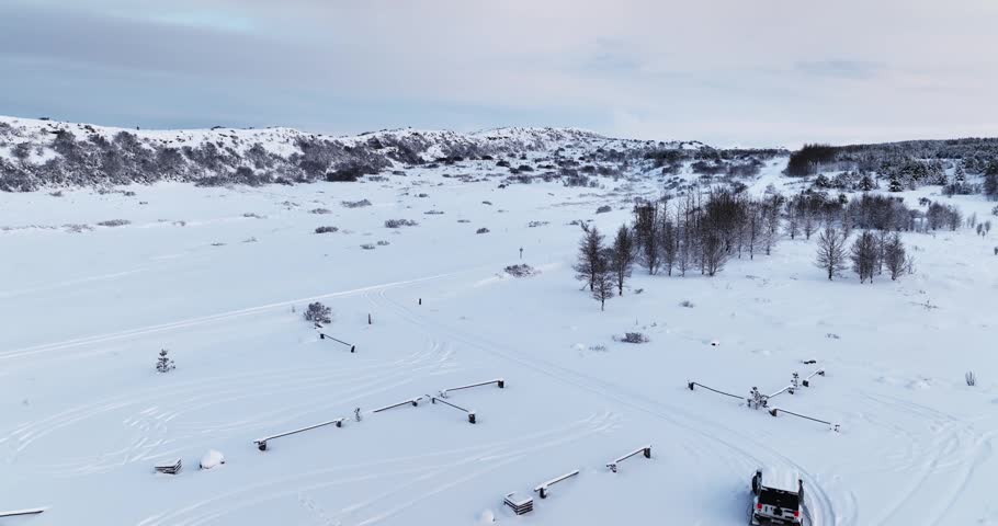 White 4x4 driving out of a snowy parking area in Heiðmörk, Iceland, with tire tracks, parking barriers, low winter hills, and frozen terrain.