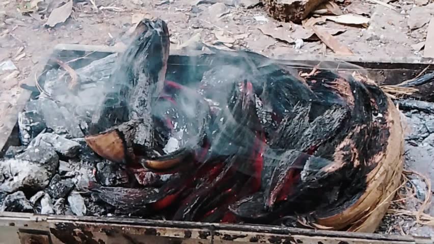 The video shows banana peels and coconut husks burning on top of glowing hot charcoal inside a small outdoor metal grill. Smoke gently rises as the husks burn over the embers.