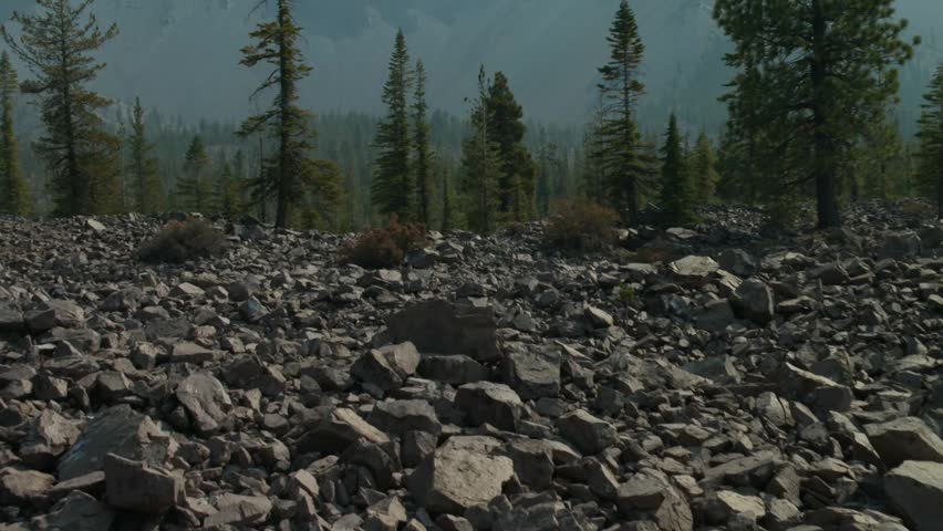 Rocky volcanic terrain with scattered conifer trees and mountain range in the background, Lassen Volcanic National Park, California, USA. Zoom out reveal shot