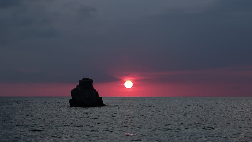Fiery crimson sunset behind a lone rock silhouette over the calm Tyrrhenian Sea, Spiaggia Michelino, Tropea, Calabria, Italy