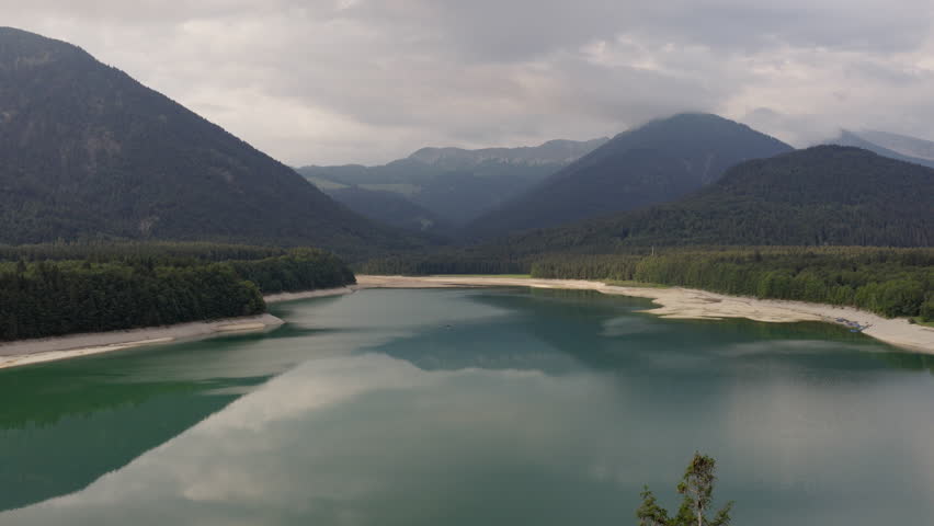 Scenic aerial view of an emerald alpine lake surrounded by forested mountains under dramatic clouds in Tyrol, Austria. Calm water reflecting the sky in a peaceful mountain landscape