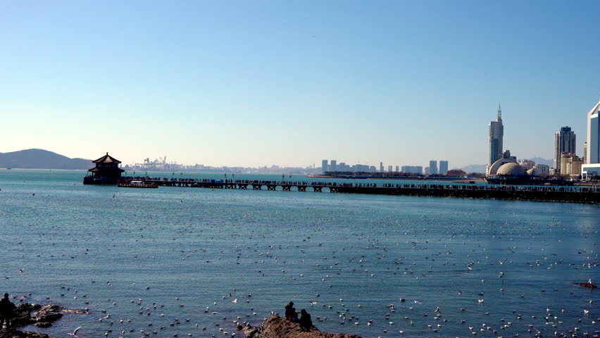 A coastal landscape showing a long pier with a traditional pavilion, modern city buildings, and calm sea under a clear blue sky.