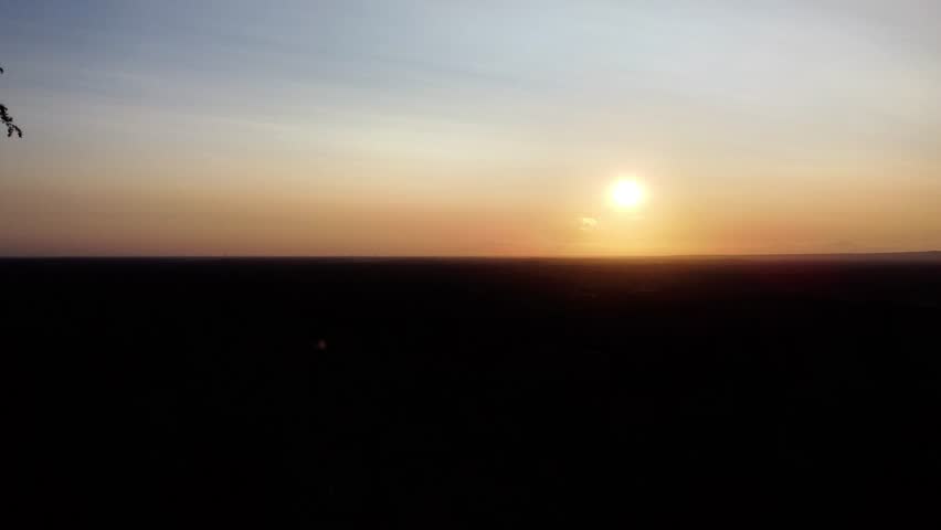 Breathtaking aerial drone footage capturing a serene summer sunset over the vast, forested landscape near Varnhem in Västra Götaland, Sweden. The bright sun hovers just above the horizon, painting the sky in beautiful shades of orange, yellow, and blue. The sprawling countryside below is cast in deep silhouette, creating a stunning contrast and a sense of tranquility. This peaceful, panoramic view showcases the natural beauty of the Swedish wilderness during the golden hour on a late summer evening.