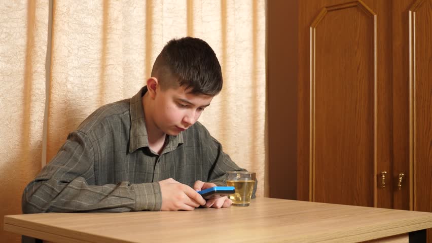 Teenager is sitting at a table, actively playing on a handheld game console, carefully controlling the game with a glass of hot tea nearby.