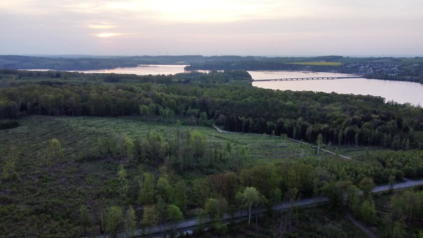 This aerial drone footage captures a stunning panoramic view of the Möhnesee reservoir in North Rhine-Westphalia, Germany. The camera glides over the dense, lush Arnsberg Forest during a tranquil spring sunset. The calm water of the large lake reflects the soft, hazy evening light. A long bridge is visible spanning a section of the reservoir, connecting the shores. A winding road meanders through the green trees and along the water