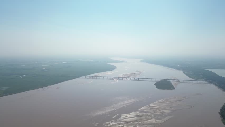 Aerial drone view of the wide Yellow River spanned by a long highway bridge. Extensive muddy sandbanks and a prominent green river island define the braided riverbed, flanked by vast green plains. The hazy summer sky highlights the grandeur of this central Chinese floodplain landscape.