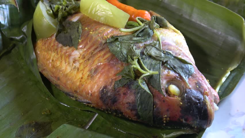 A close-up video of a tilapia head that has been cooked after being boiled. This steamed red tilapia dish in green banana leaves is served warm with pre-coated spices. Several vegetables are also added for aroma and flavor, such as chopped basil leaves, red chilies, and sliced ​​green tomatoes to enhance the taste. It is served warm on a clean white plate.