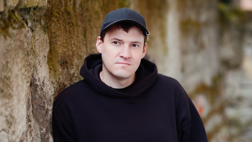 Man in black hoodie and cap leaning against rustic stone wall. Casual portrait. Urban street style outdoor session.