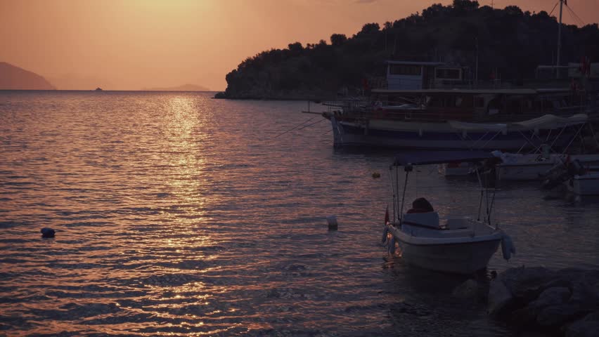peaceful marine bay during a warm golden sunset, with several small boats and yachts moored near the rocky shoreline. The setting sun casts a brilliant, shimmering orange reflection across the rippling sea surface, illuminating the water and the silhouettes of hills in the distance. The composition captures the tranquil evening atmosphere by the coast, highlighting the interplay of light and water. This image documents a serene seaside view at dusk, offering a scenic visual for travel content, coastal lifestyle projects, nautical themes, or nature stock footage emphasizing quiet harbor moments and evening tranquility