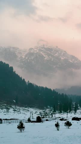 4K Vertical view of snowfall in beautiful mountain valley during sunset at Kashmir, India. Orange sunset clouds in sky and snow covered mountains at Aru Valley during winter season. Dramatic nature landscape.